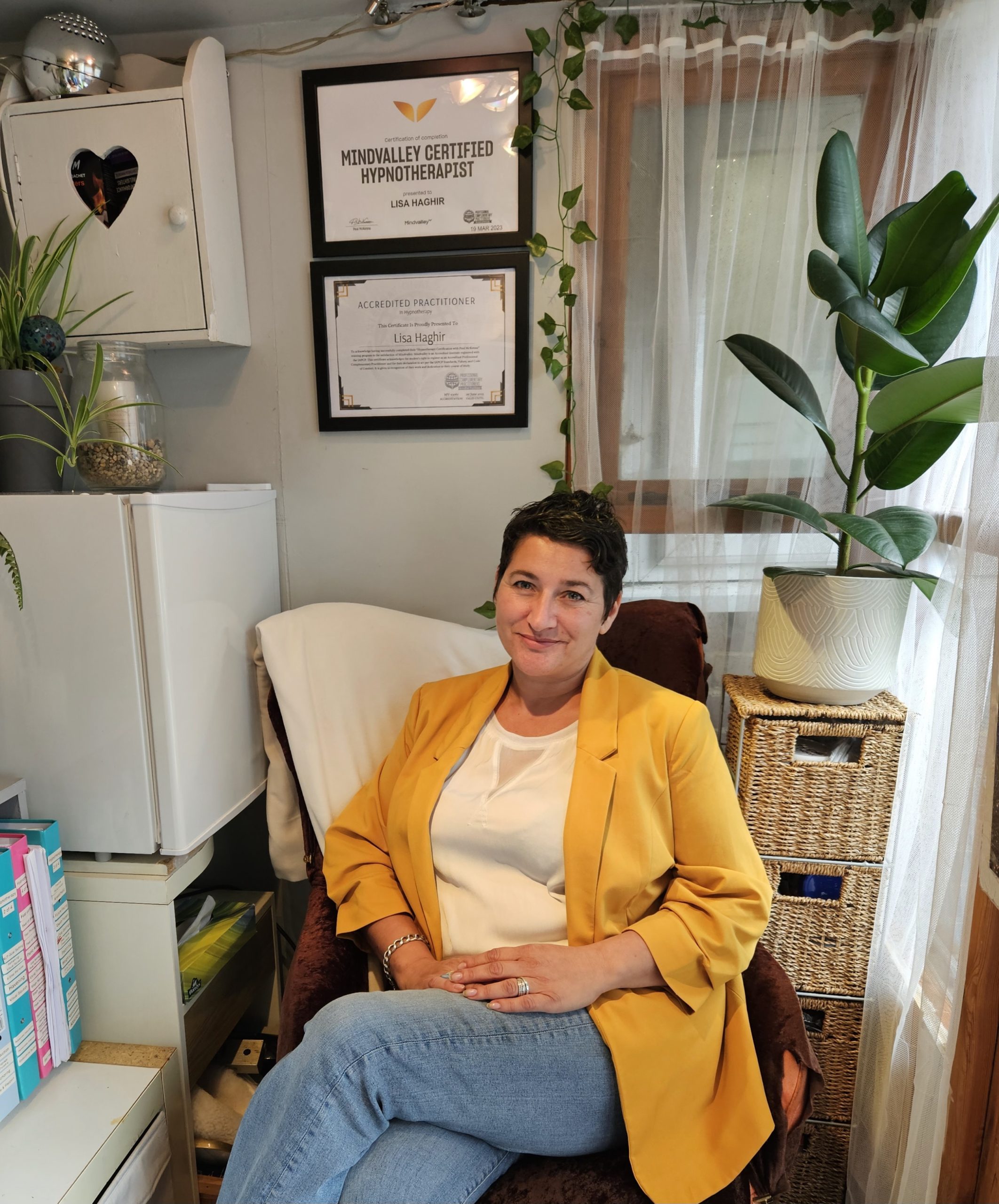Lisa a Hypnotherapist wearing a bright mustard jacket sitting in her home office with some plants, and you can see her certificates on the wall in the background